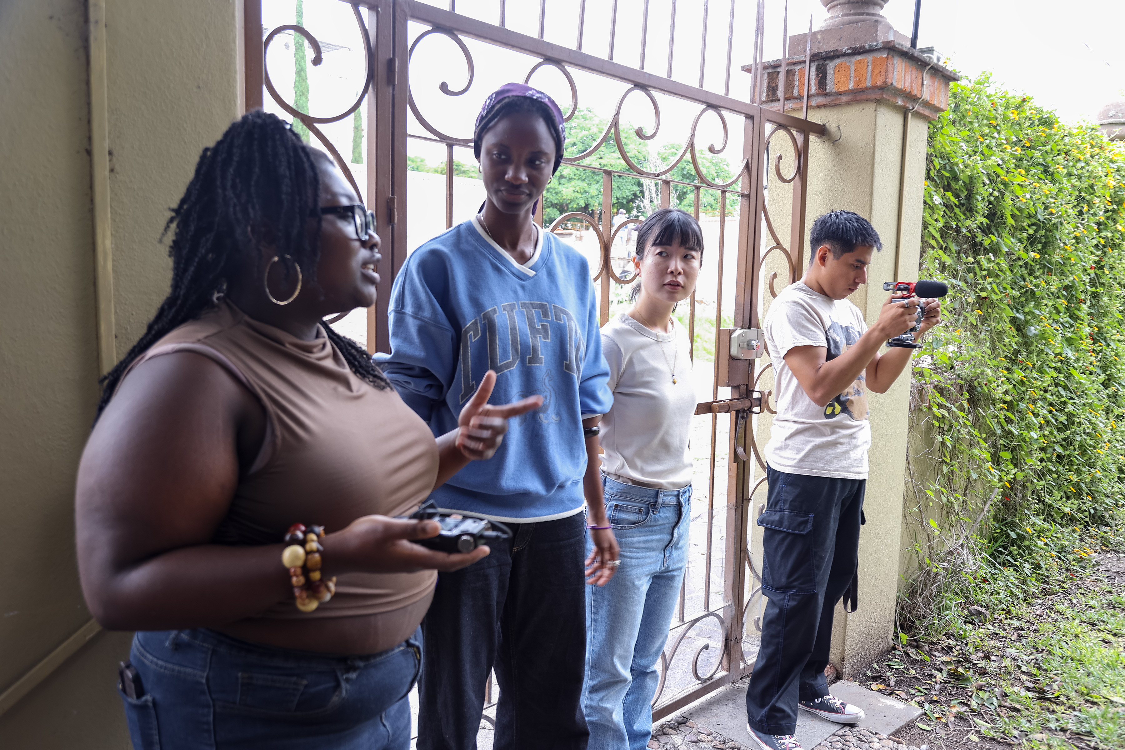 Four student documentary filmmakers stand outside a wrought-iron gate, one holding a camera with a shotgun mic, mid-discussion — illustrating the responsibility of ethical documentary filmmaking before the camera rolls.