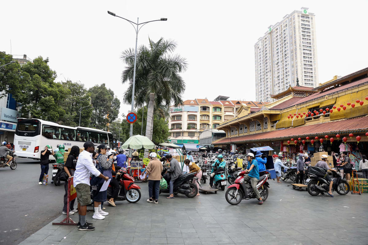 Busy street market in Ho Chi Minh City, Vietnam, with vendors, motorcycles, and pedestrians in front of a traditional market building.