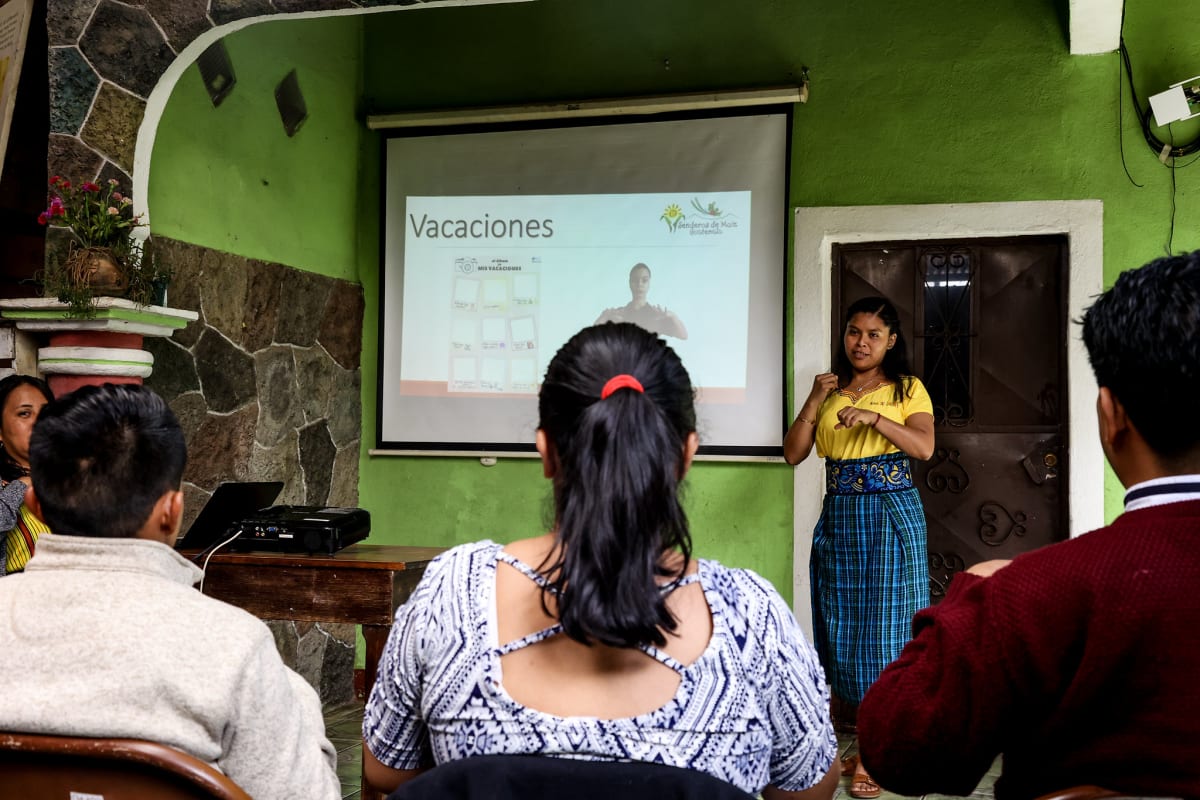 A staff member at Alma de Colores leads an after-hours sign language class for colleagues in Guatemala, with a projected presentation visible behind her.