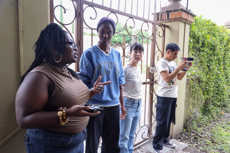 Four student documentary filmmakers stand outside a wrought-iron gate, one holding a camera with a shotgun mic, mid-discussion — illustrating the responsibility of ethical documentary filmmaking before the camera rolls.