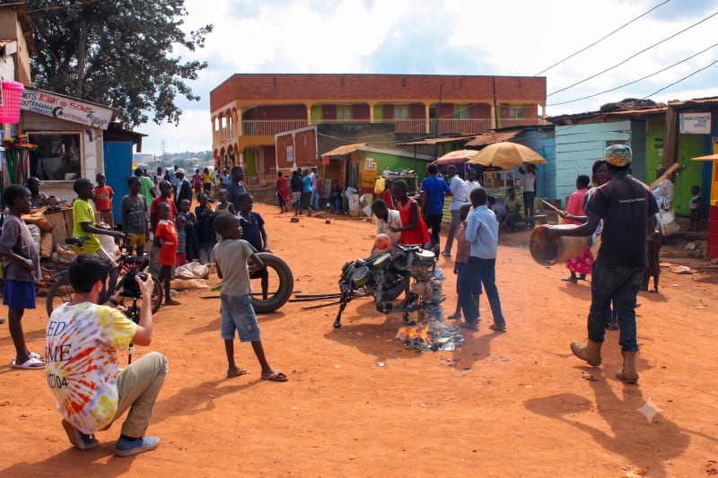 A documentary filmmaker crouches in the foreground shooting a street scene in Kampala, Uganda, where a crowd of children and community members has gathered around a metal sculpture — an example of ethical documentary filmmaking in the field.
