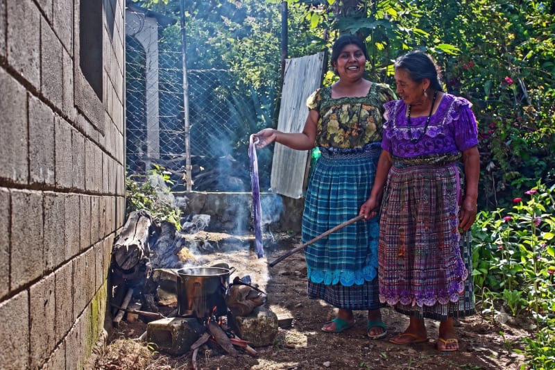 Two Tz'utujil Maya women from Asociación Lema' stand beside a wood fire, lifting freshly dyed fabric from a pot — a scene from responsible documentary storytelling in San Juan la Laguna, Guatemala.