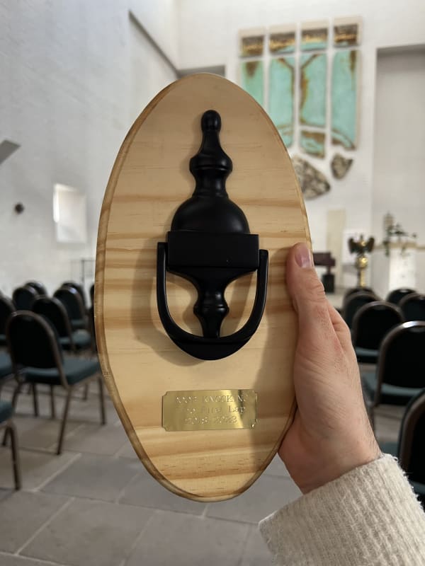 A homemade door knocking trophy with a door knocker mounted on a wooden board