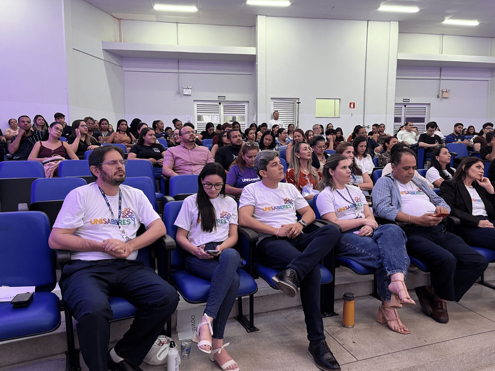 Equipe do UniSaberes com camisetas do evento sentada na plateia do auditório da UnirG