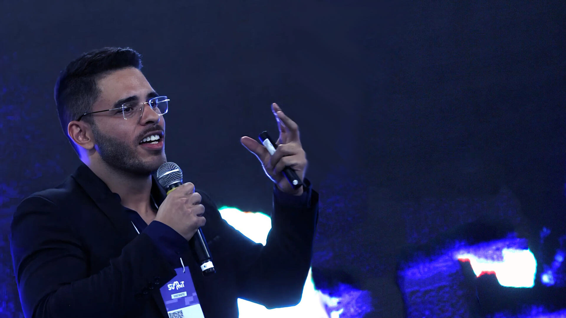 Luiz Carvalho palestrando em evento, segurando um microfone e um controle remoto, com expressão envolvente. Ele usa óculos e veste um blazer escuro, destacando-se contra um fundo azul iluminado por luzes difusas. Um crachá de identificação pendurado em seu pescoço sugere participação em um evento tecnológico