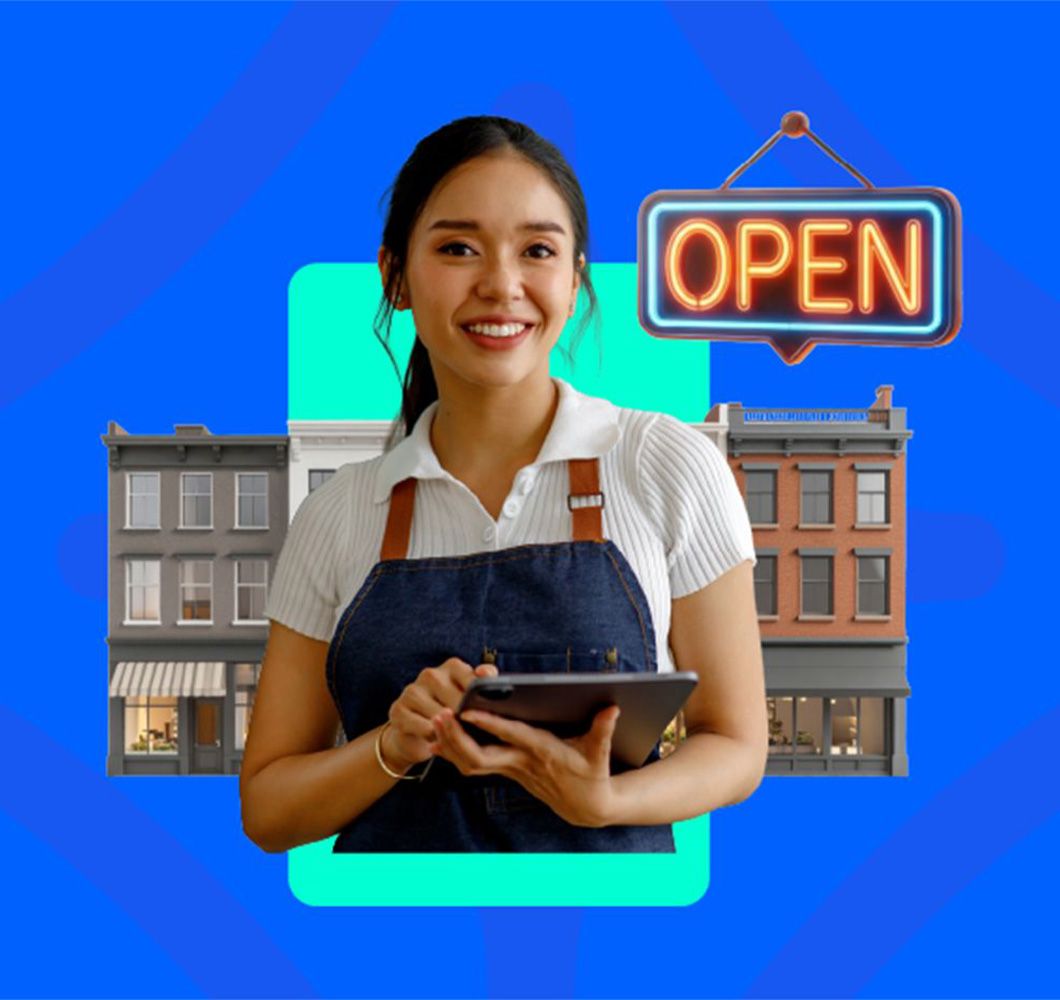 Smiling small business owner in a denim apron holding a tablet in front of Main Street storefronts and a neon Open sign