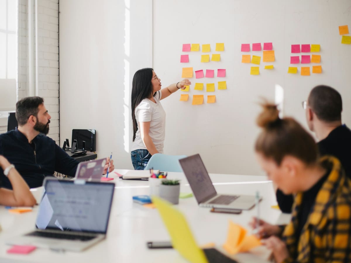 A trainer leading a workshop at a wall of sticky notes while a team works on laptops