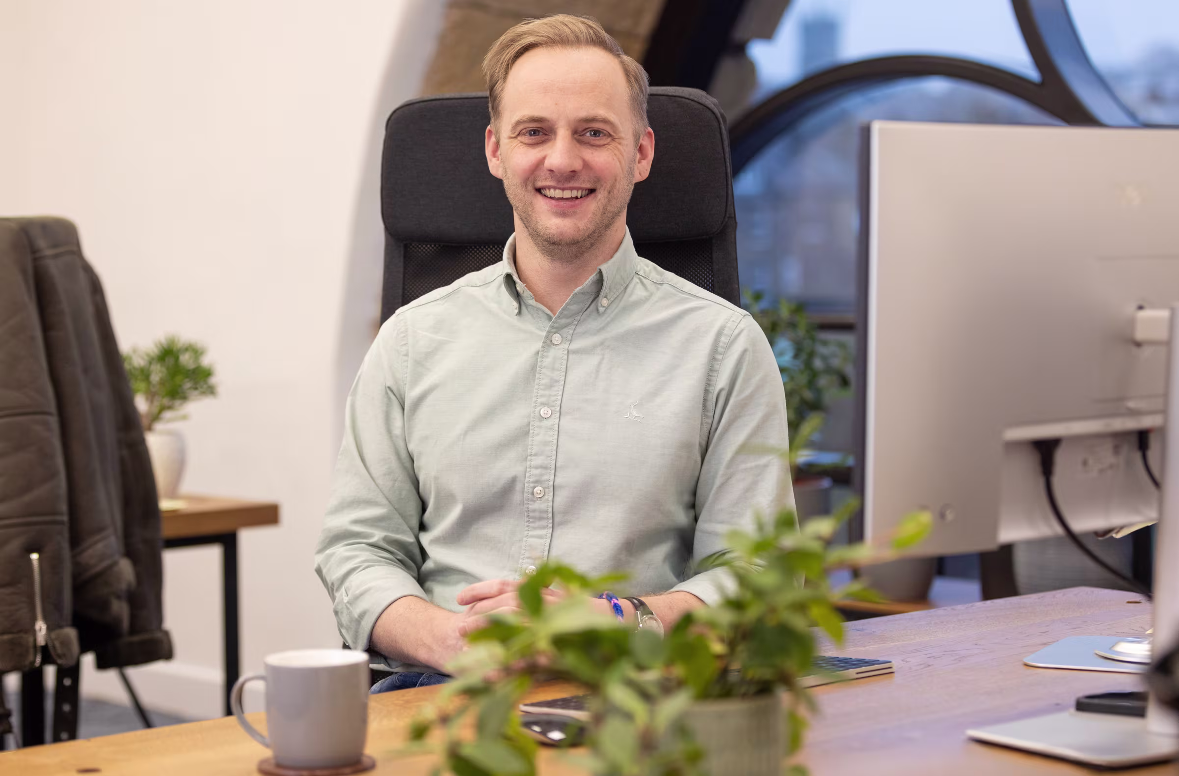 A professional headshot of Chris Nicholl, a man with light brown hair, smiling while sitting at a modern wooden desk in a bright office. He is wearing a light green button-down shirt, with a computer monitor and potted plants in the background.