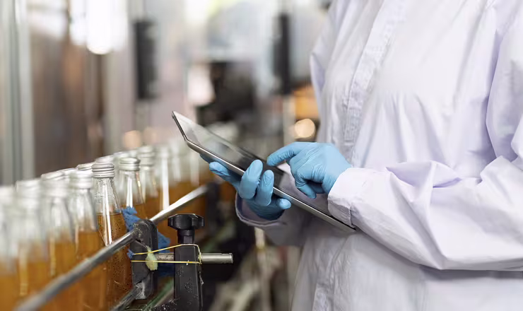 Laboratory technician wearing gloves and a lab coat inspecting bottles using a tablet.