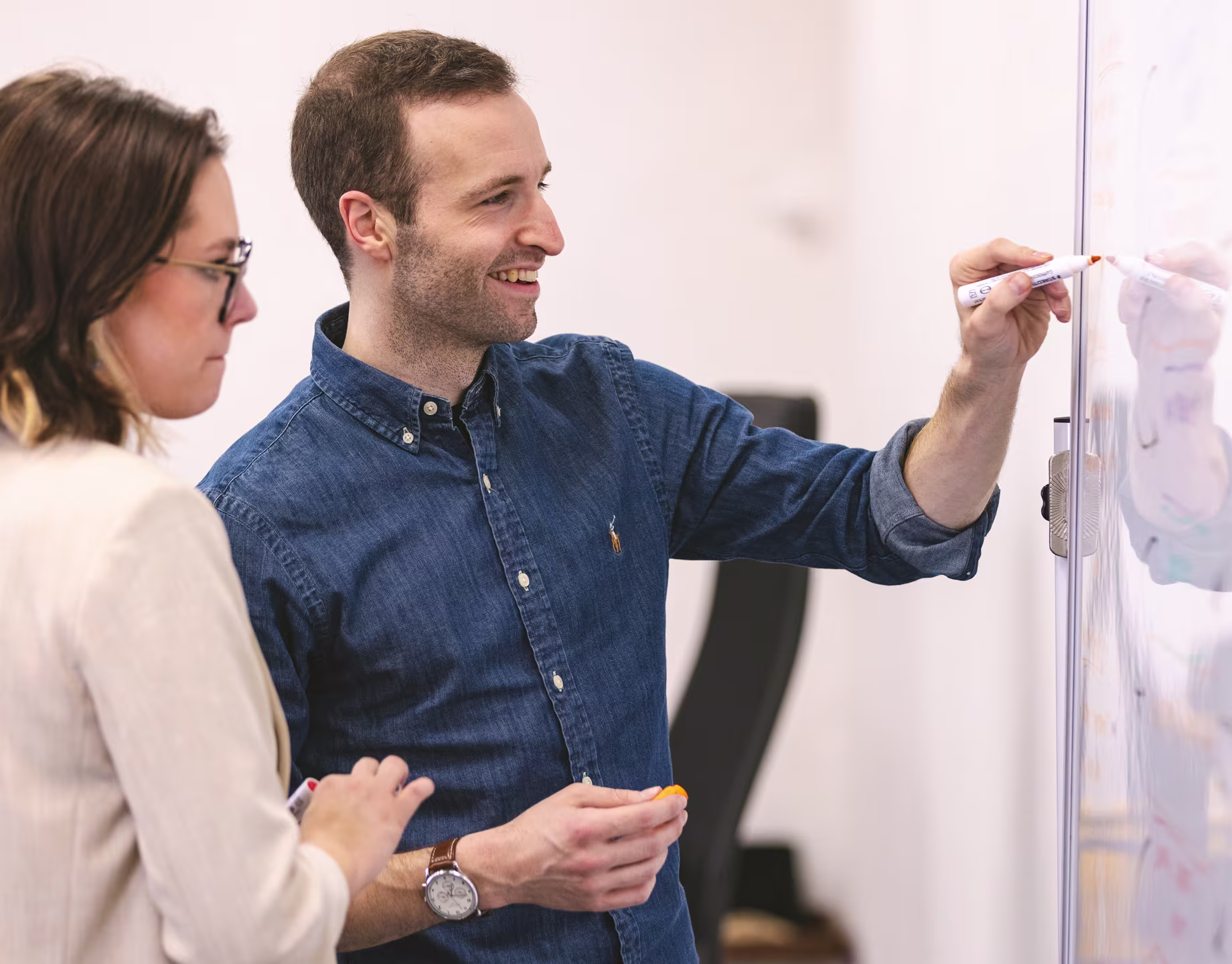 A male consultant writing on a whiteboard while explaining a technical concept to a female colleague