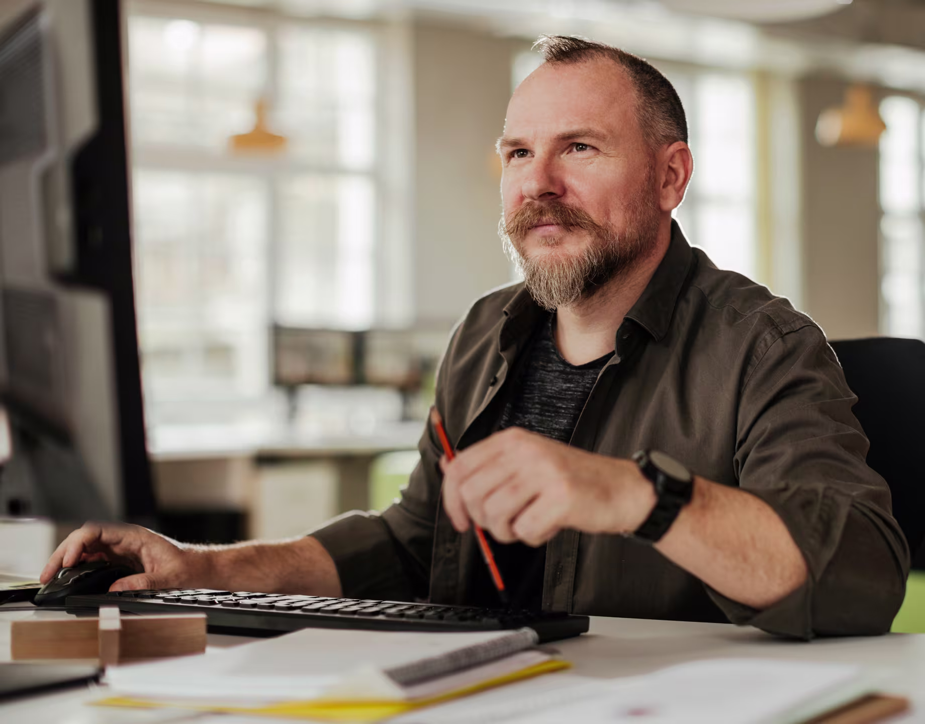 Middle-aged man with a beard working at a computer in a bright modern office, holding a pencil and looking thoughtfully at the screen.