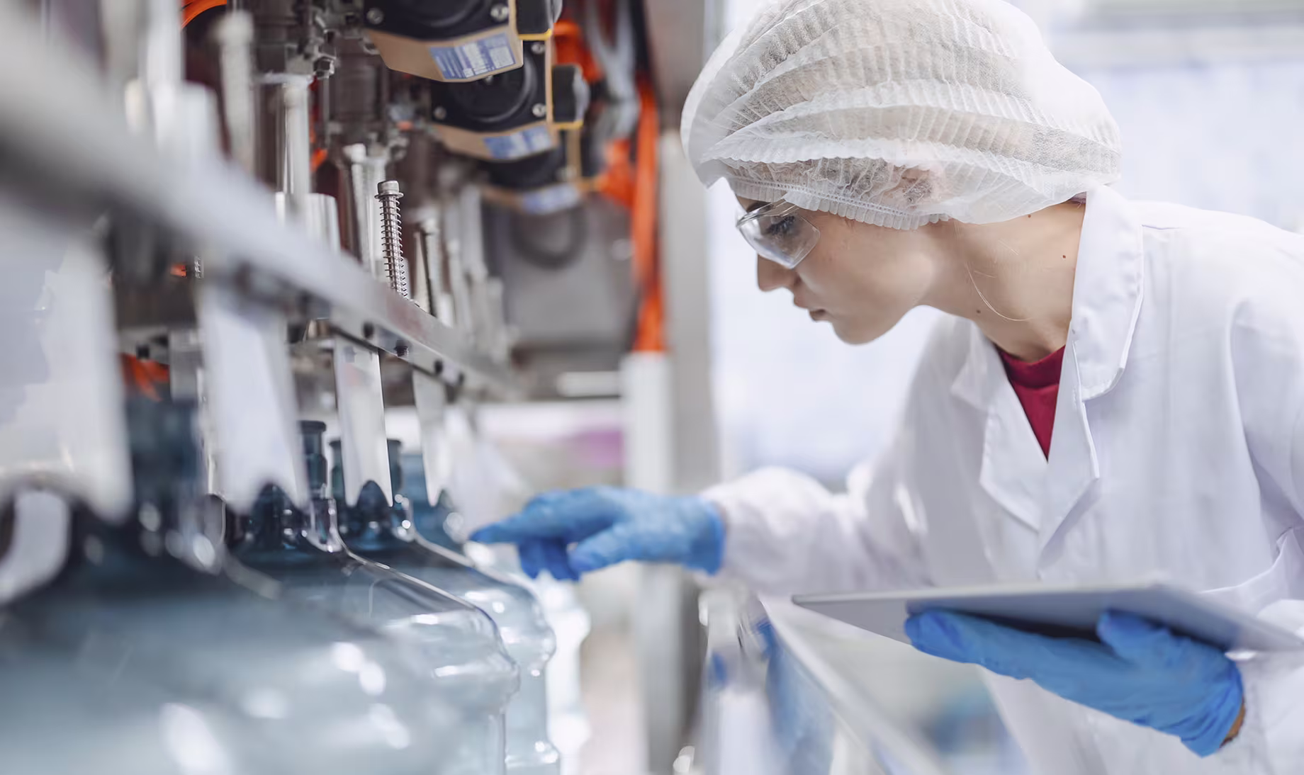 A person in protective gear inspects a machine with large bottles in an industrial setting, holding a tablet.