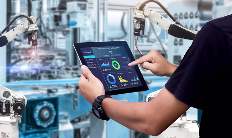 Person operates a tablet showing data analytics dashboard in a modern industrial setting with robotic arms and machinery in the background.