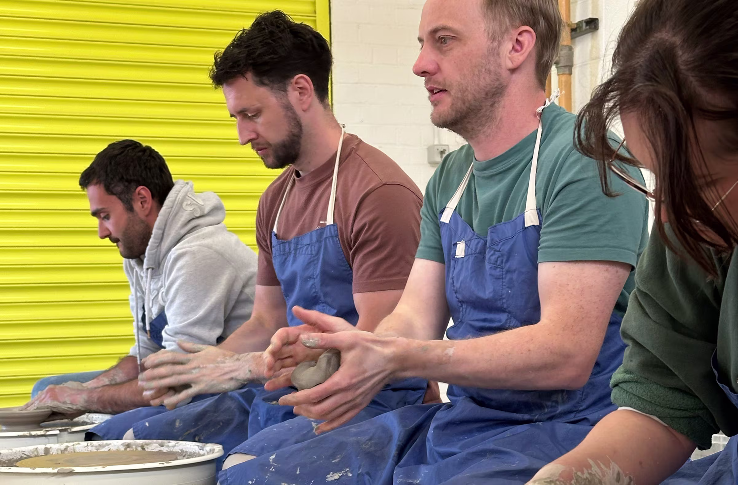 A group of people, including Chris Nicholl, participating in a pottery workshop. They are wearing blue aprons and sitting in a row, focusing on shaping clay on pottery wheels in a studio with a yellow shutter door in the background.