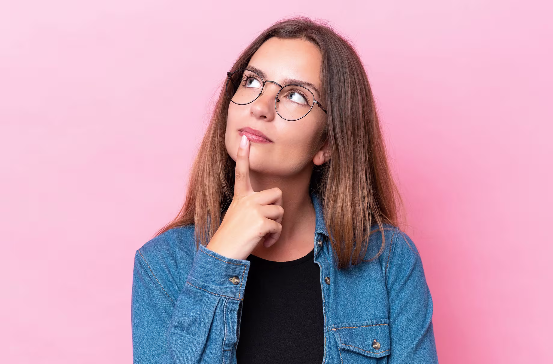 Young woman with glasses wearing a denim jacket, looking pensive against a soft pink background.