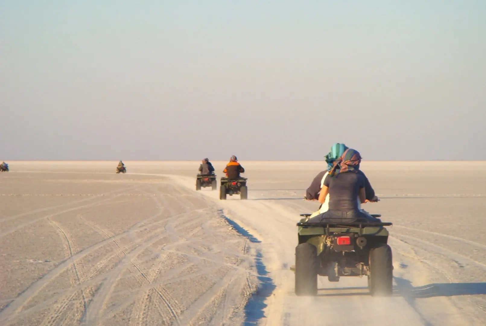 Four quads lined up one after the other on a dusty desert trail, with people riding them through the vast expanse of Makgadikgadi Pan.