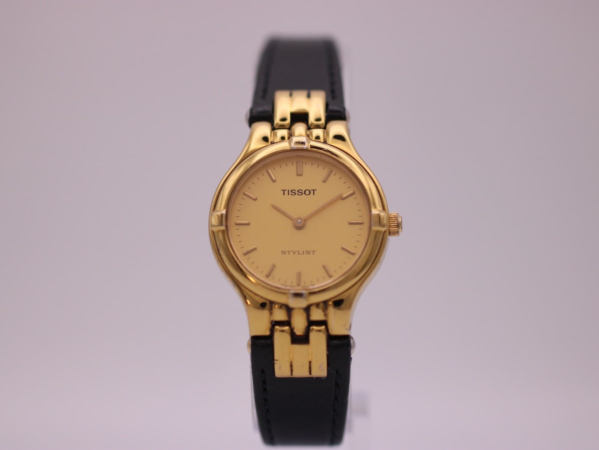 Front view of the Tissot Stylist watch, showing the gold dial, gold-tone case, and black leather strap.