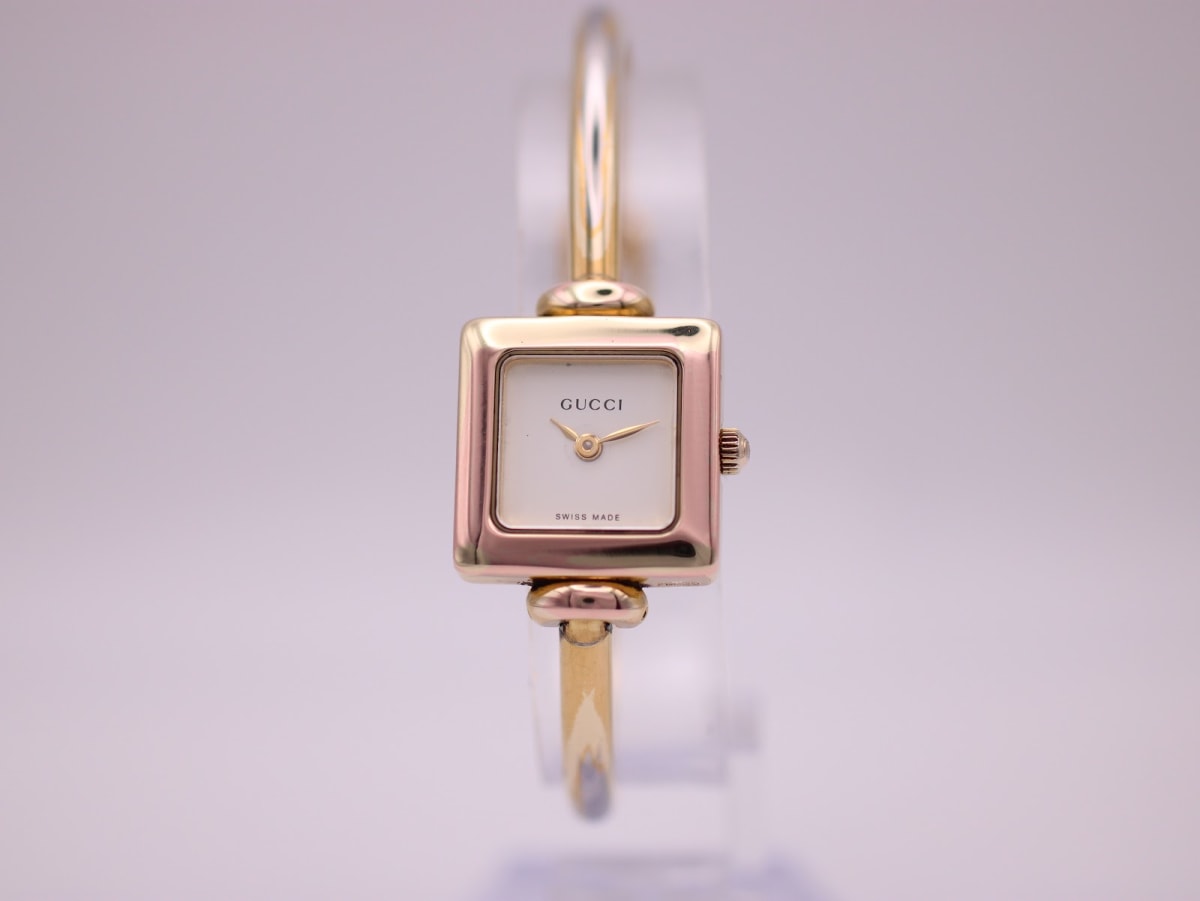 Front view of the Gucci 1900L watch with a square gold-tone case and cream dial, on a clear stand against a white background.