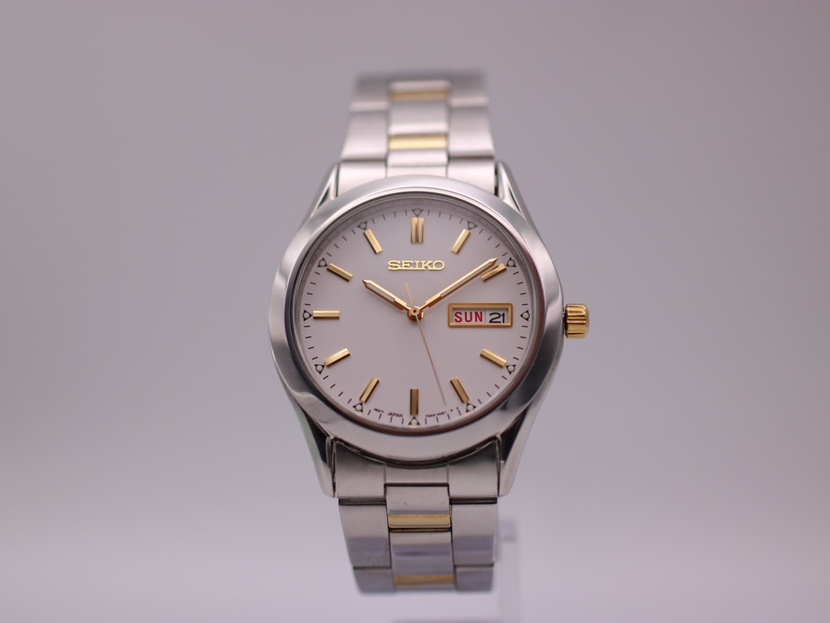 Front view of the Seiko 7N43-9070 watch with white dial, gold baton indices, and two-tone stainless steel bracelet on a grey background.