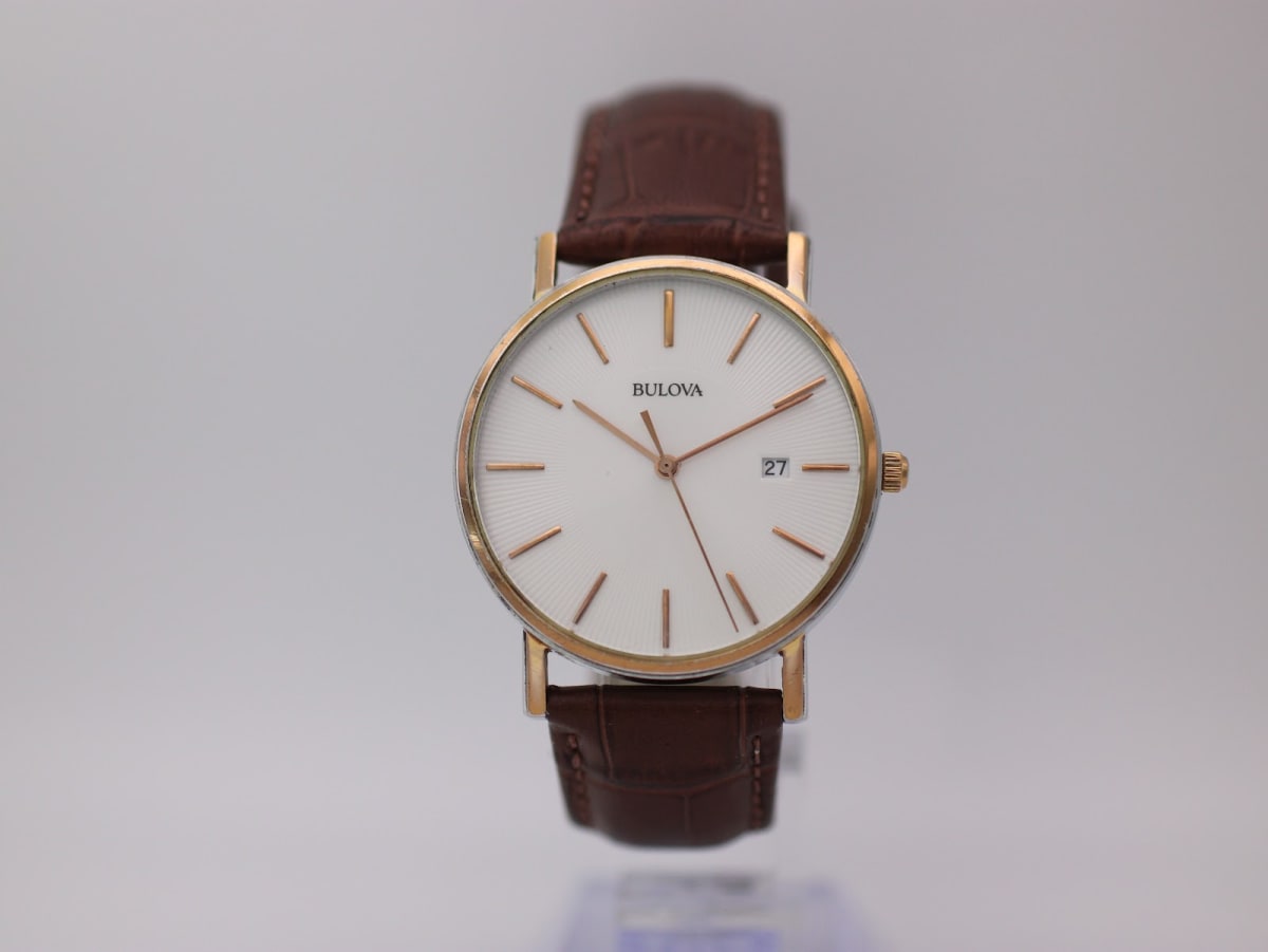 Front view of Bulova C4331132 two-tone watch with white textured dial, rose gold-tone baton indices, and date window, on a brown leather strap.