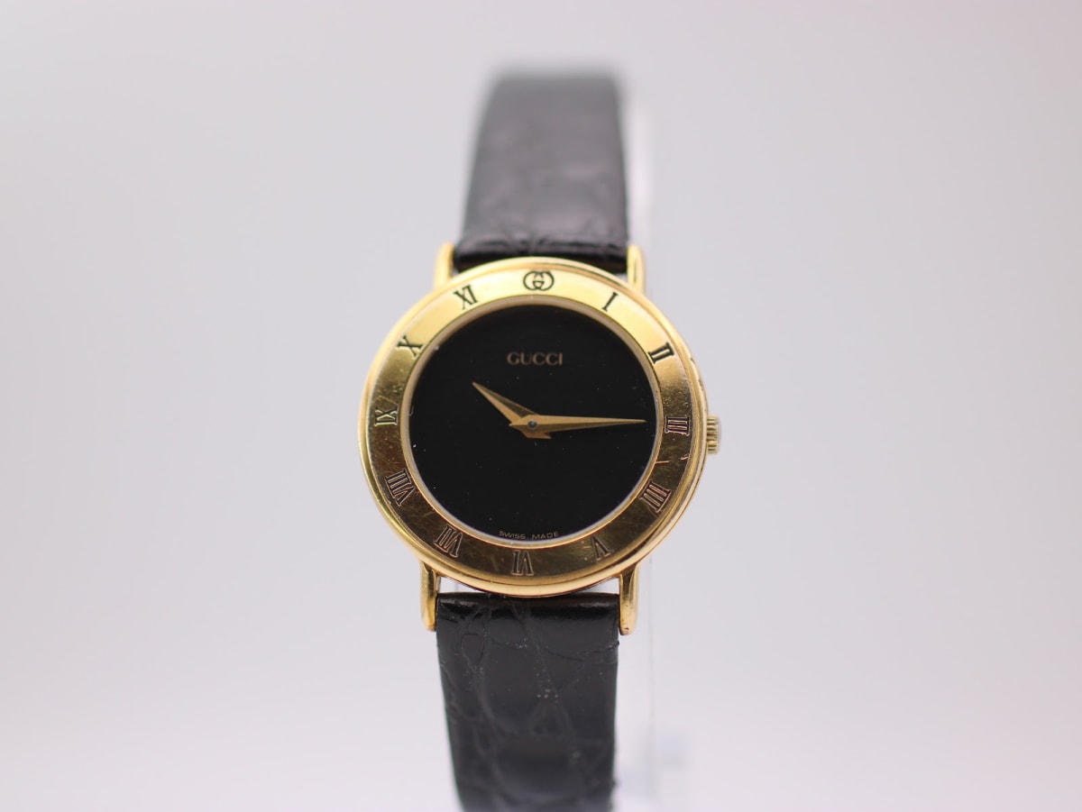 Front view of the gold-plated Gucci 3000.2.L quartz watch with a black dial and roman numeral bezel on a white stand.