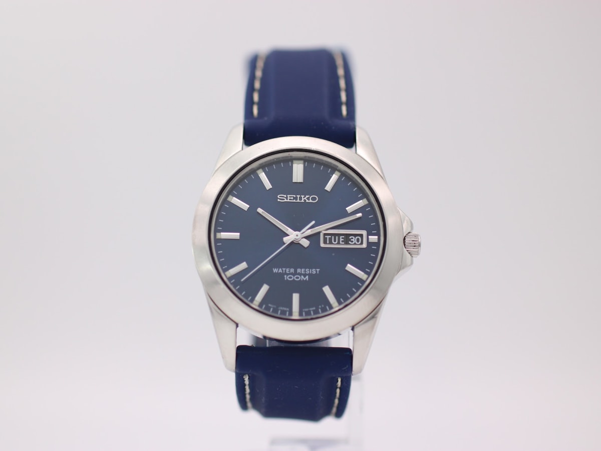 Front view of the Seiko V743-9A20 watch, silver case, blue dial with day-date, and navy rubber strap, on a clear stand.