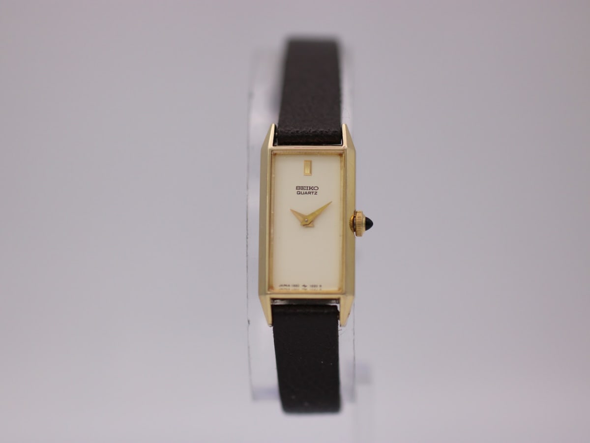 Front view of the gold-tone rectangular Seiko watch with cream dial and brown leather strap on a watch stand.