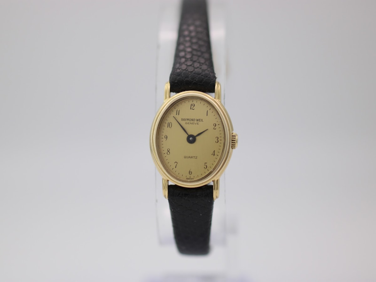 Front view of the Raymond Weil 5717 watch, showing the round gold-toned case, champagne dial with black numerals, and black lizard-grain leather strap on a clear stand.