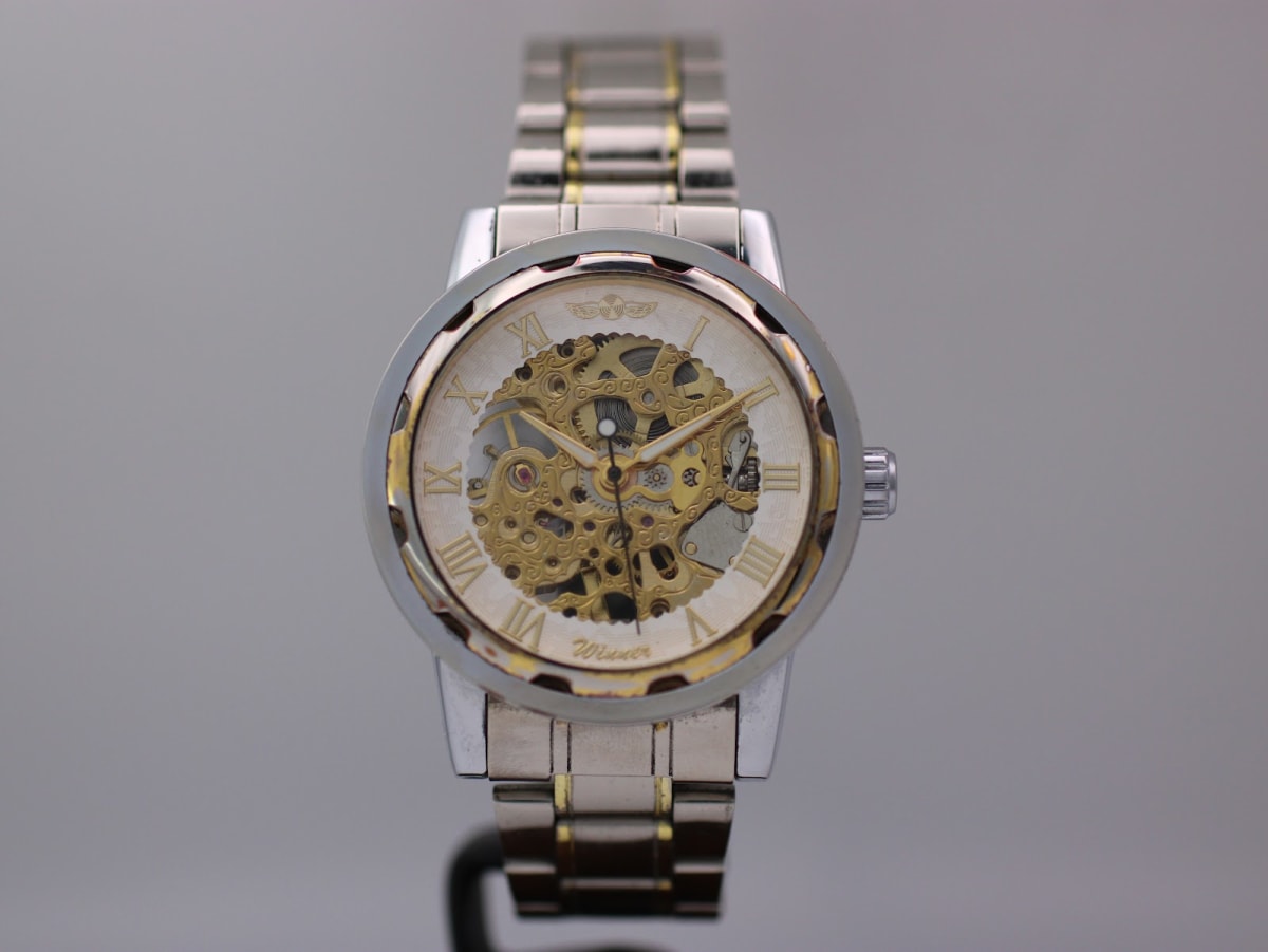 Front view of the two-tone Winner skeleton watch with silver dial and gold Roman numerals, on a display stand.