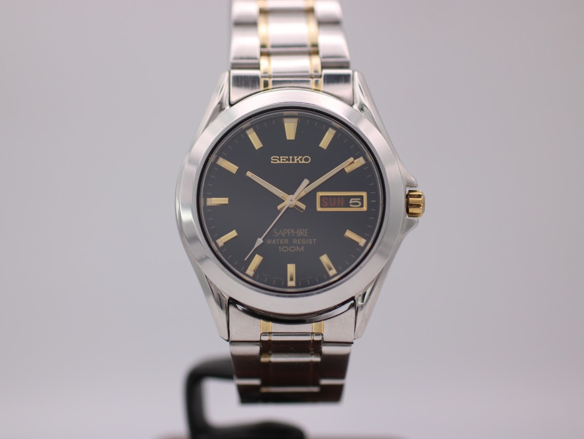 Front view of Seiko Sapphire Day-Date watch with black dial, gold baton indices, and two-tone steel bracelet.