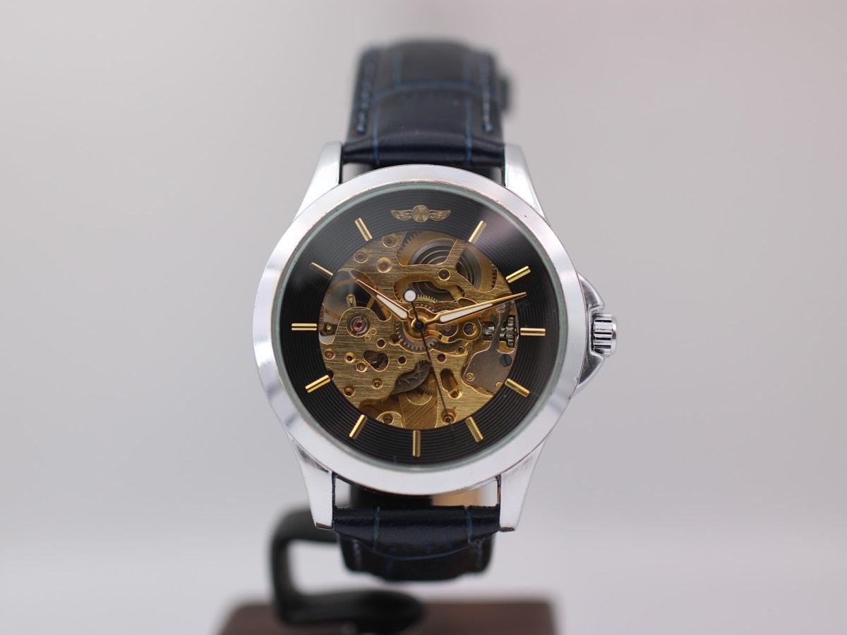 Front view of generic skeleton automatic watch with a black dial and gold-tone movement on a stand.
