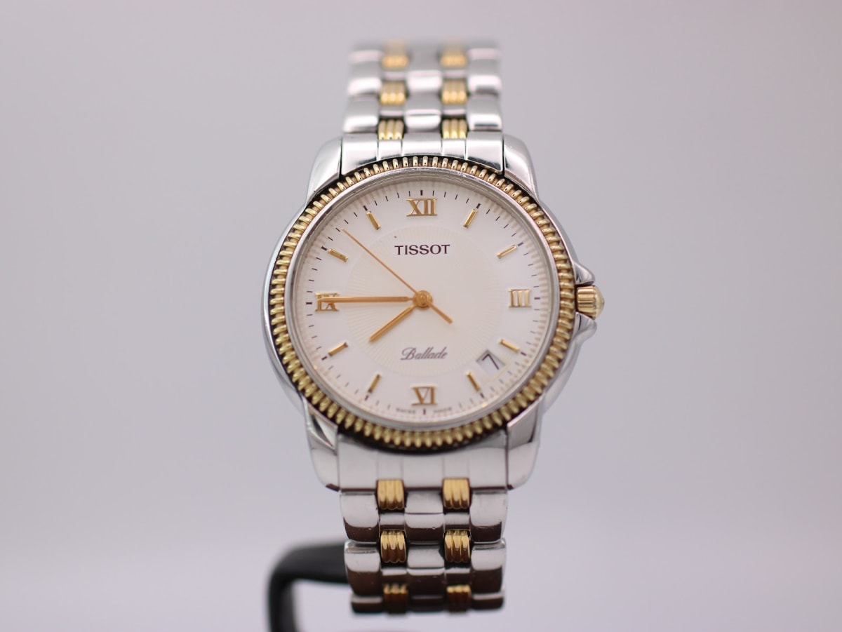 Front view of the Tissot Ballade two-tone watch, showcasing the cream dial with gold Roman numerals and hands on a watch stand.