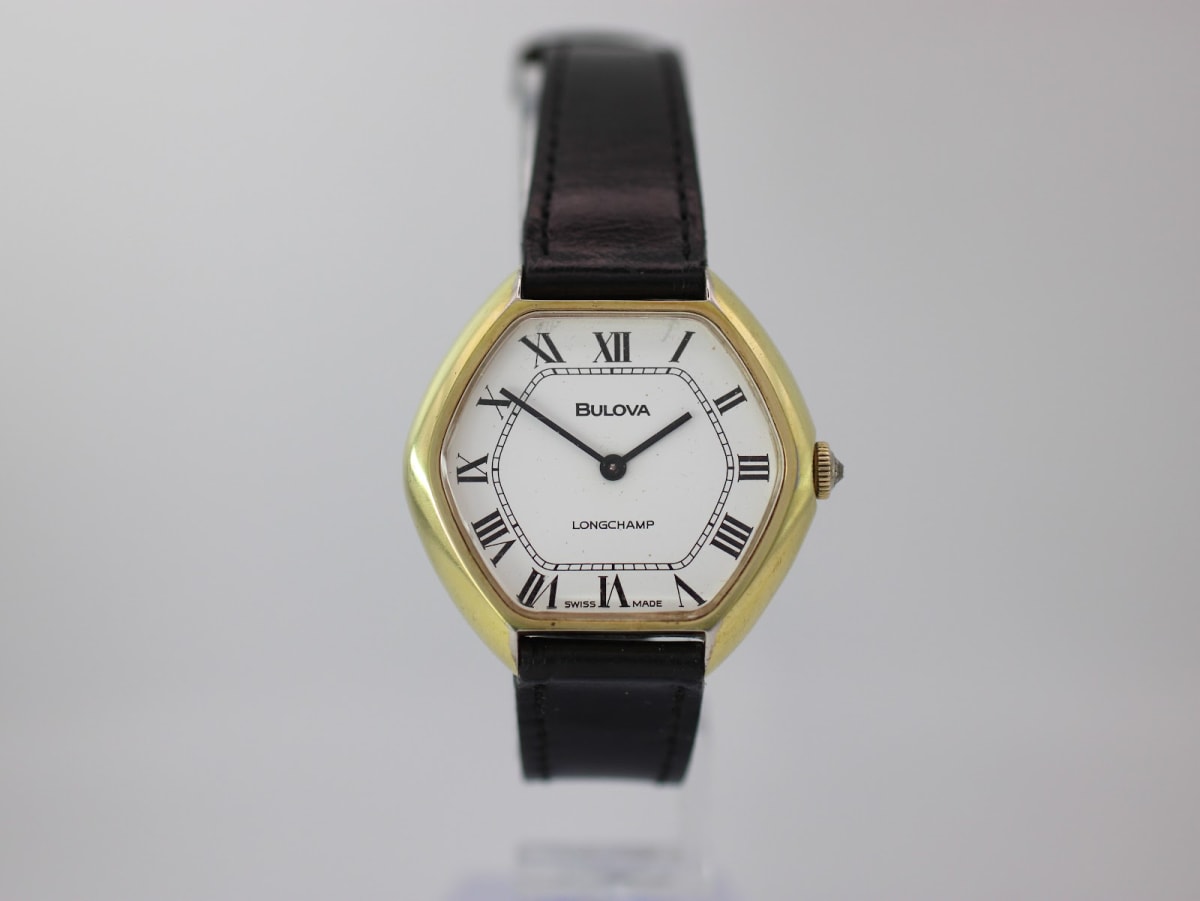 Front view of the Bulova Longchamp watch on a clear stand, showing the octagonal gold case, white Roman numeral dial, and black leather strap.