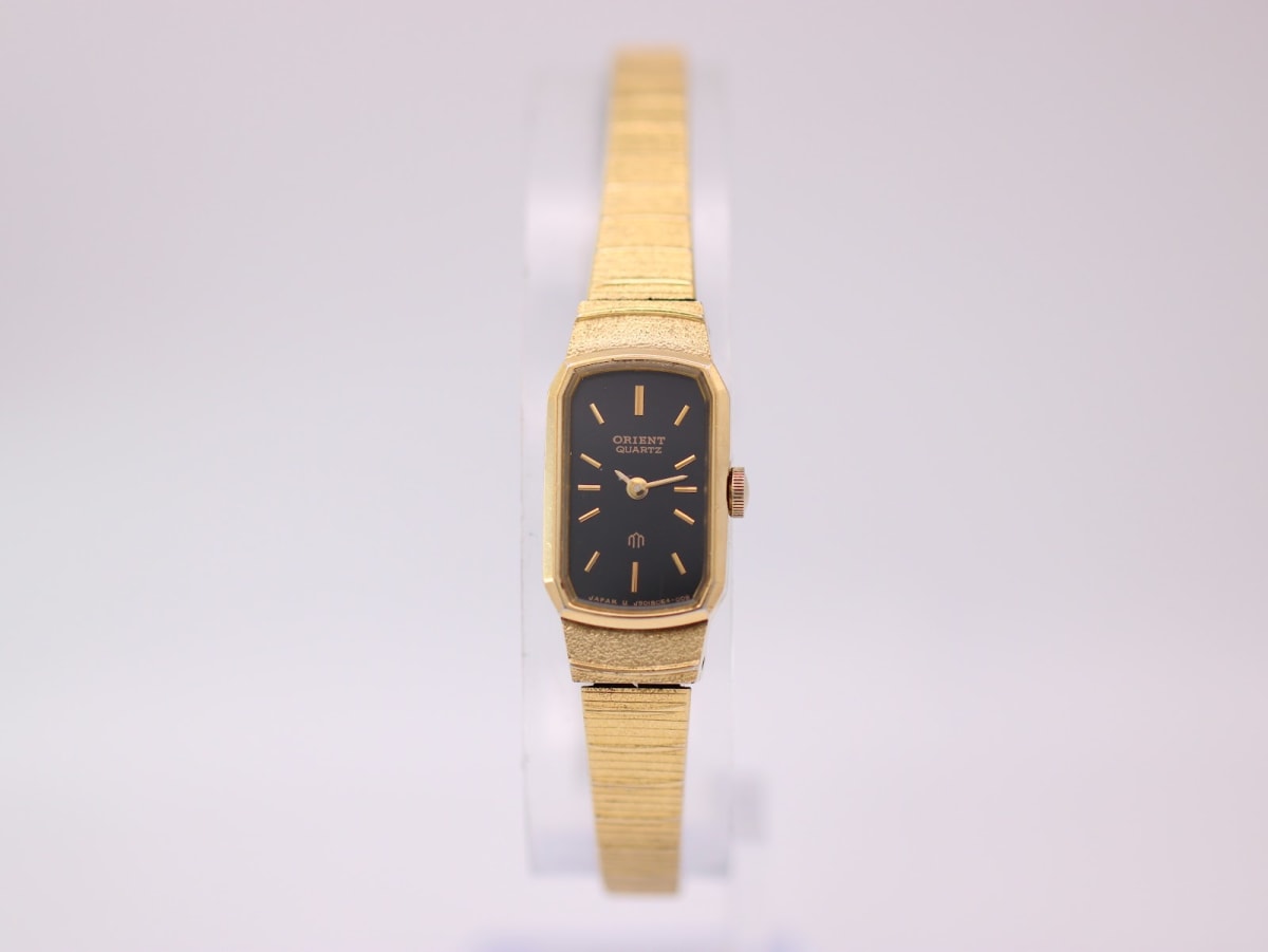 Front view of the gold-tone Orient quartz watch on a clear stand, showing the black rectangular dial and integrated bracelet.