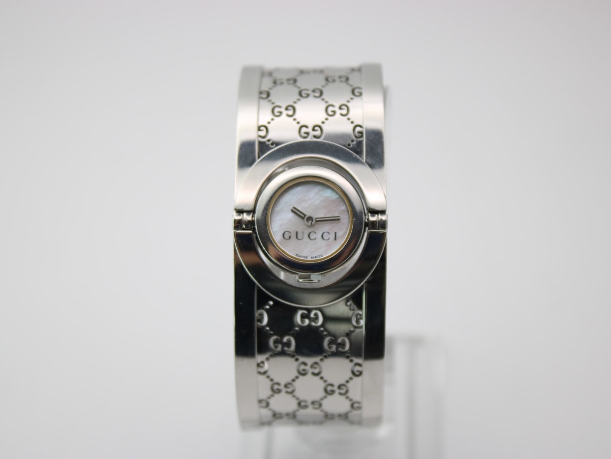 Front view of the Gucci 112 Twirl watch, showcasing its mother-of-pearl dial and engraved stainless steel bangle on a display stand.