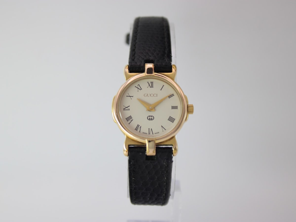 Front view of the Gucci 3400 L watch on a clear display stand, showing the cream dial with roman numerals and black leather strap.