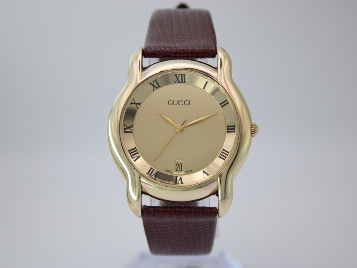 Front view of gold tone Gucci 5100 M watch with gold dial and brown lizard grain leather strap, on a clear display stand.