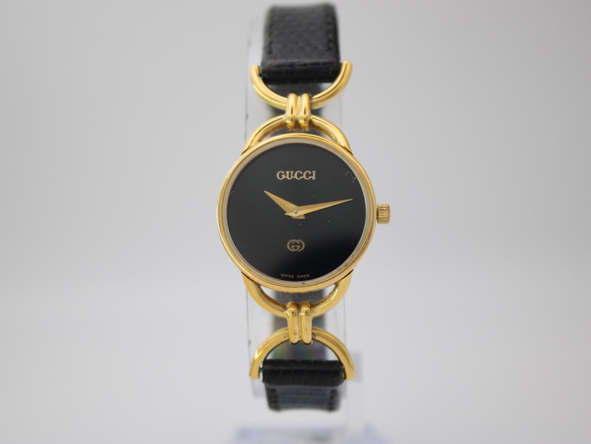 Front view of the Gucci 6000L watch on a clear display stand, showing the black dial, gold-tone case, and black leather strap.