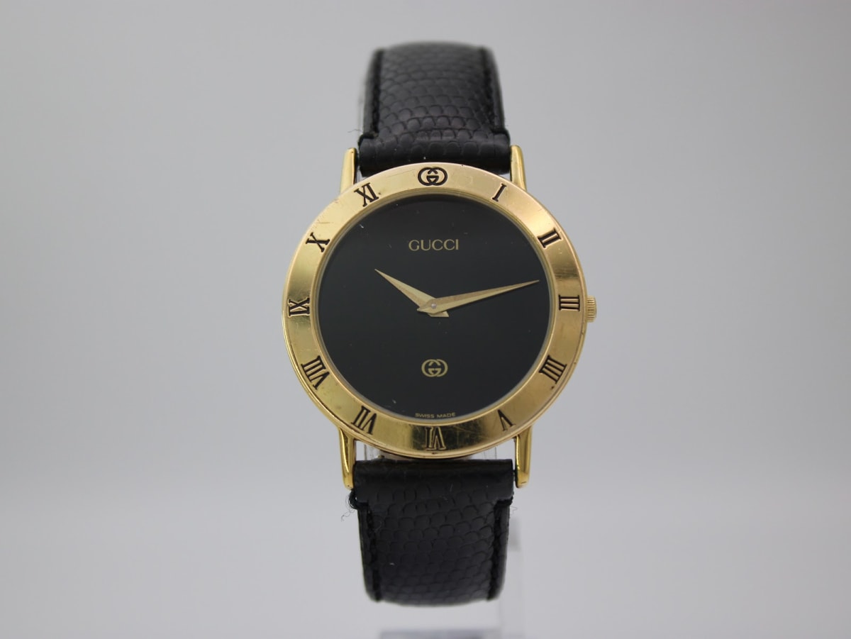 Front view of the Gucci 3000 M watch on a clear stand, showing its black dial, gold-tone case, and black lizard-effect leather strap.
