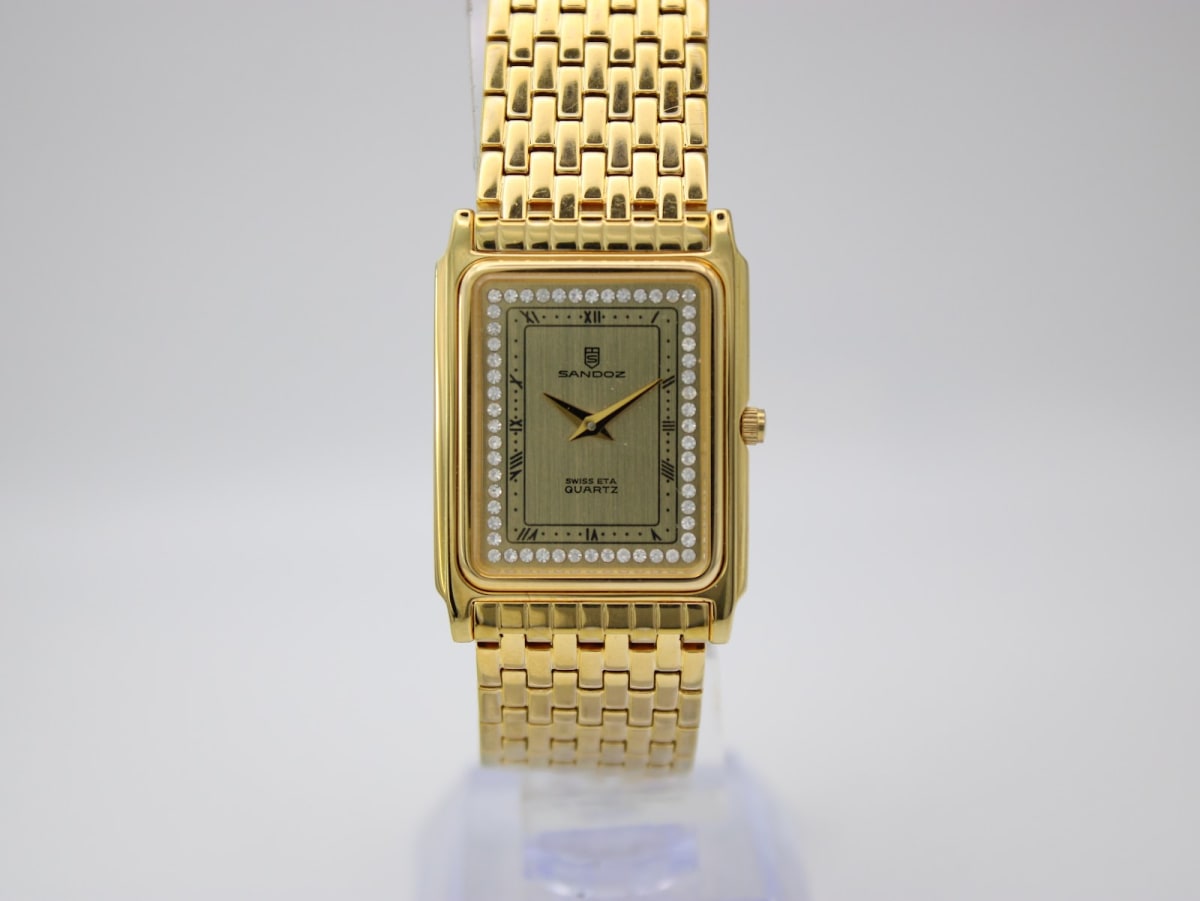 Front view of Sandoz rectangular gold-tone watch with champagne dial and rhinestone bezel on a clear stand.