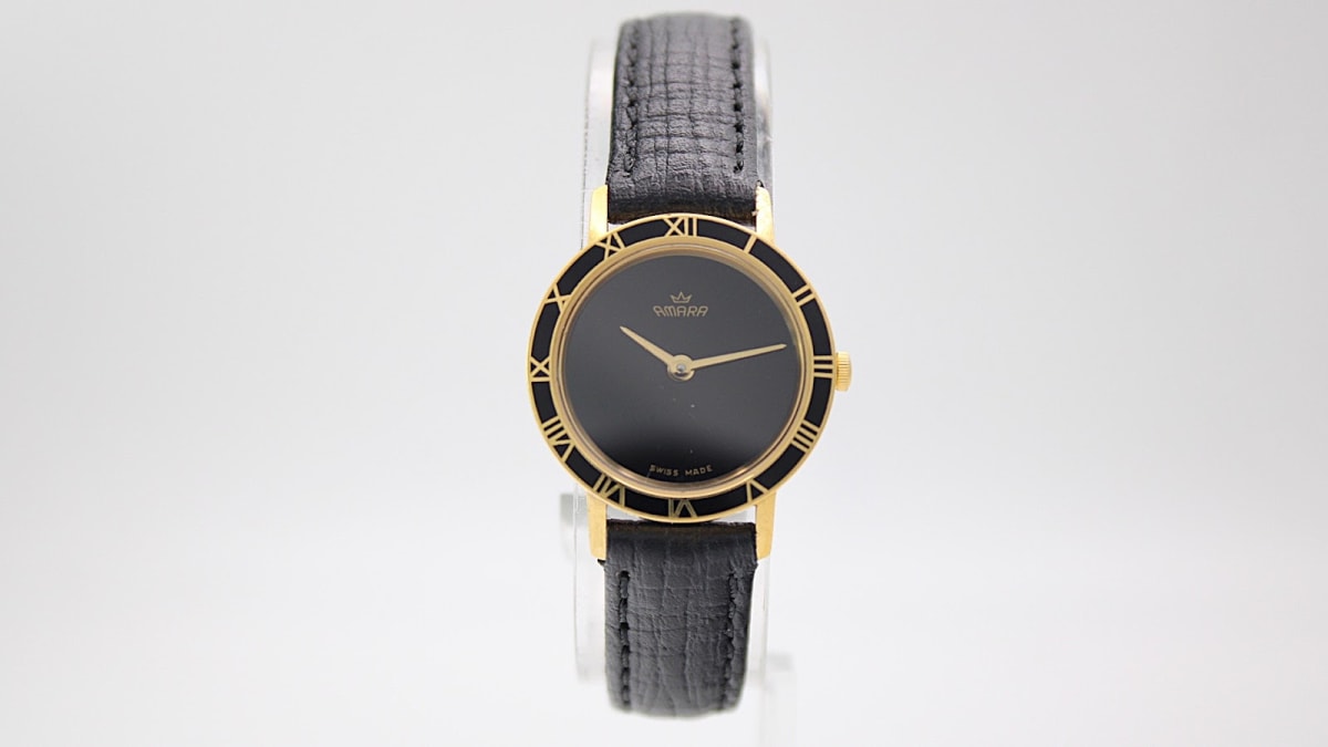 Front view of the Amara watch with a gold-tone case and a plain black dial, featuring gold Roman numerals on the bezel and a black leather strap.