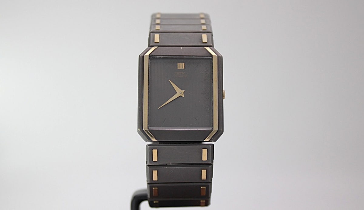 Front view of Seiko 5470-7000 rectangular two-tone watch with black dial and gold-tone hands on a stand.