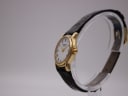Side view of the Raymond Weil watch showing the gold case, crown, and black leather strap on a clear watch stand.