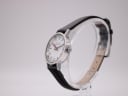 Angled side view of the Mondaine watch showing the silver case profile and black leather strap on a clear stand.