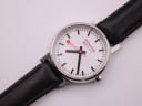 Close-up front view of the Mondaine white dial with clear black baton hour markers and the iconic red seconds hand, partially showing the black leather strap.