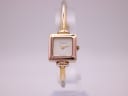 Front view of the Gucci 1900L watch with a square gold-tone case and cream dial, on a clear stand against a white background.