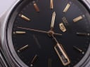 Close-up of the black dial showing gold baton indices, gold hands, Seiko logo, and day-date window.