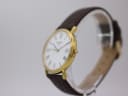 Angled side view of Tissot watch showing gold case, crown, and brown strap on a clear display stand.