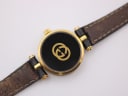 Close-up back view of the Gucci 2000L watch case, featuring the prominent gold-toned interlocking G logo against a black background.