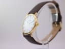 Angled side view of the Raymond Weil watch showing the gold case, crown, and brown leather strap on a display stand.